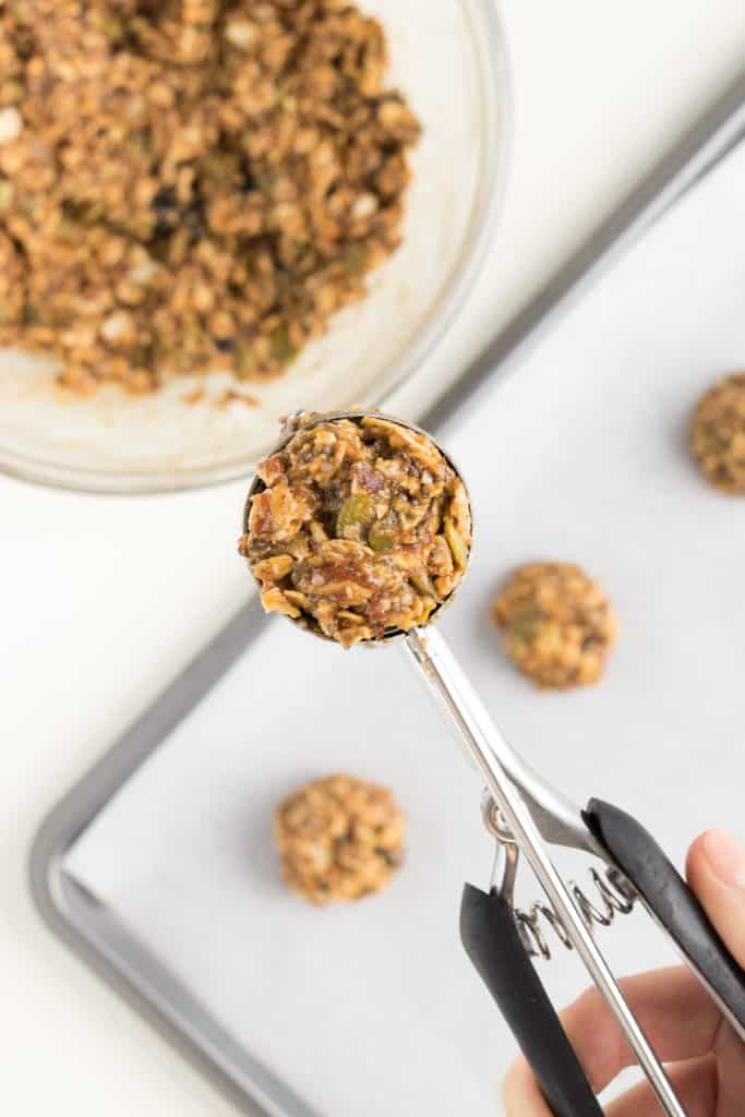 hand holding a metal cookie scoop filled with breakfast cookie dough in front of a metal baking sheet and glass bowl