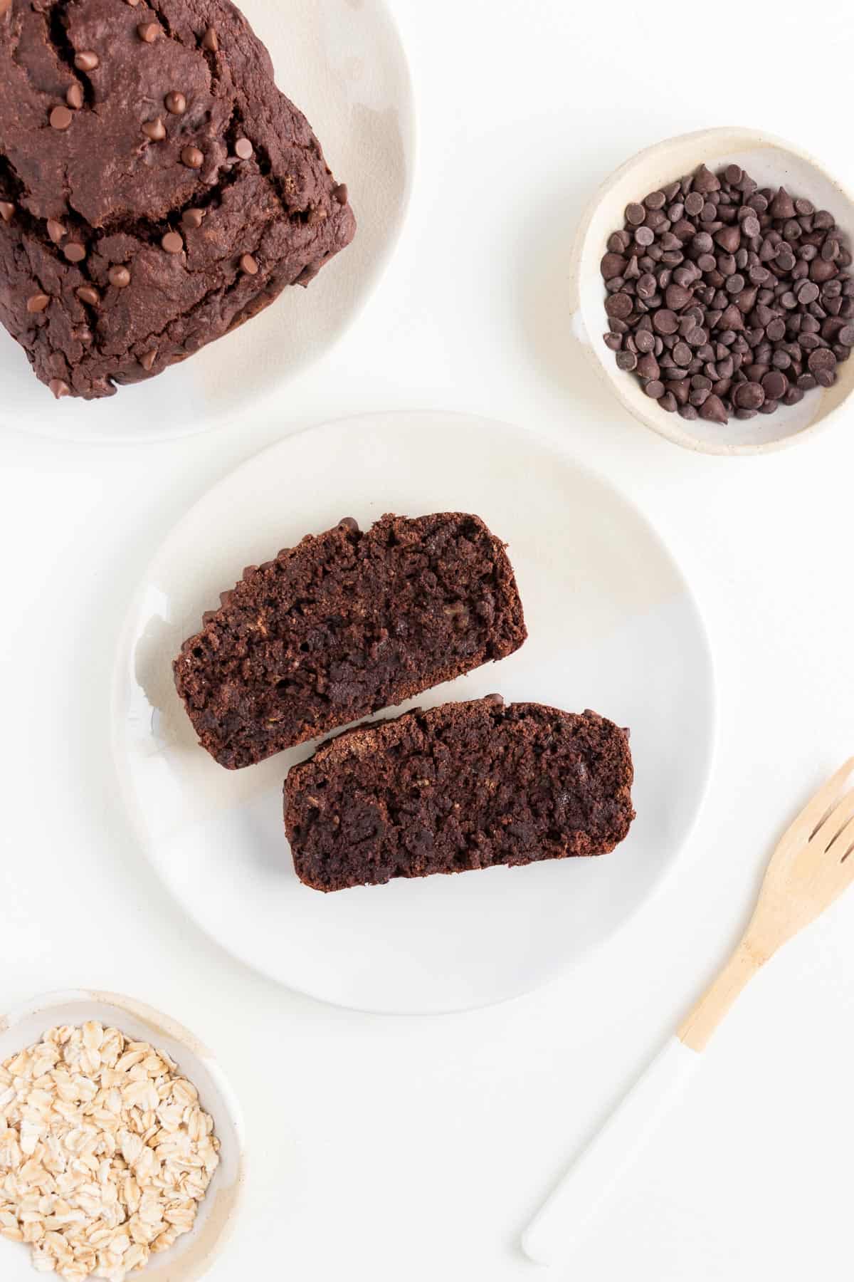 two slices of vegan gluten-free chocolate banana bread on a white plate surrounded by a small bowl of chocolate chips and a small bowl of rolled oats