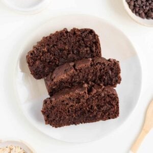 three slices of chocolate banana bread on a white plate surrounded by a wooden fork, a cream mug filled with almond milk, and a small bowl of rolled oats