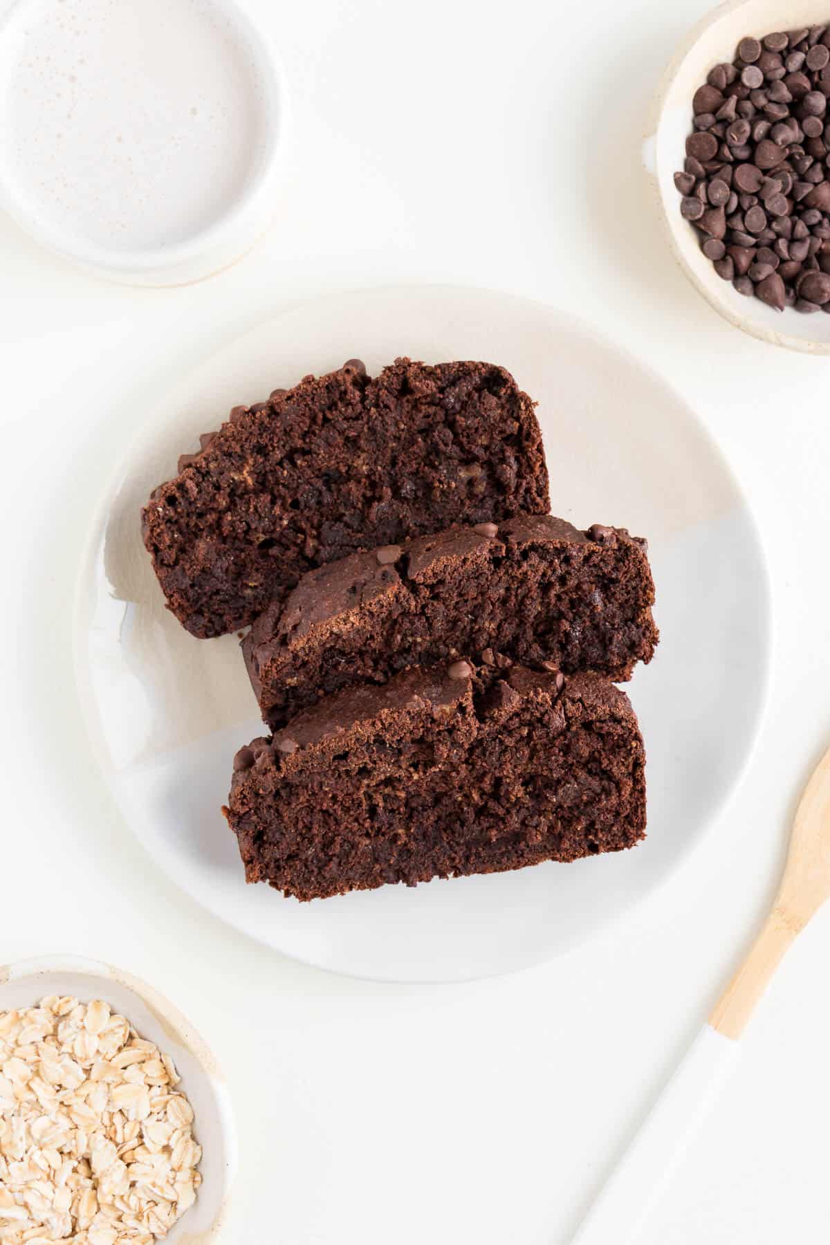 three slices of chocolate banana bread on a white plate surrounded by a wooden fork, a cream mug filled with almond milk, and a small bowl of rolled oats