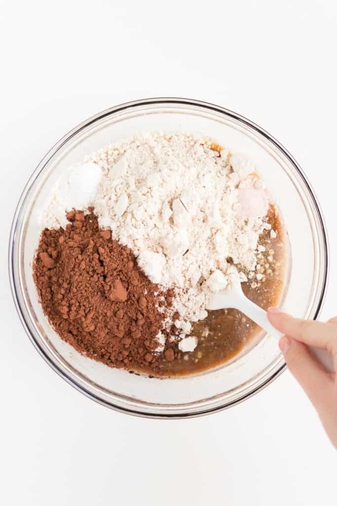 hand holding a white spatula mixing four and cocoa powder in a glass bowl