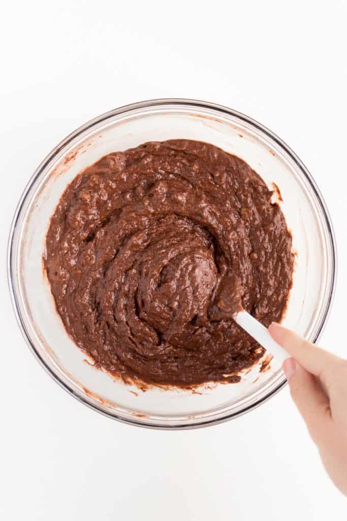 hand holding a white spatula mixing chocolate banana bread batter in a glass bowl