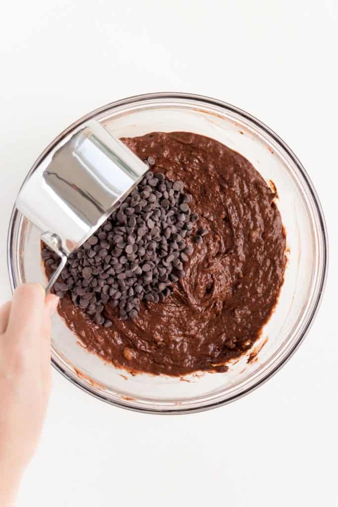 hand holding a metal measuring cup pouring chocolate chips into chocolate batter