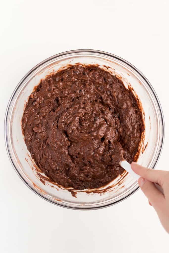 hand holding a white spatula mixing chocolate batter inside a glass bowl