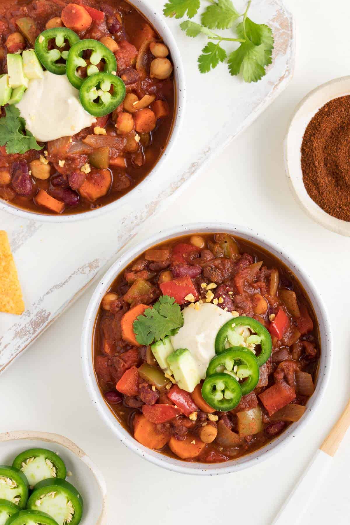 two white ceramic bowls filled with vegan three bean chili alongside a wooden spoon, bowl of jalapeños, and bowl of chili powder