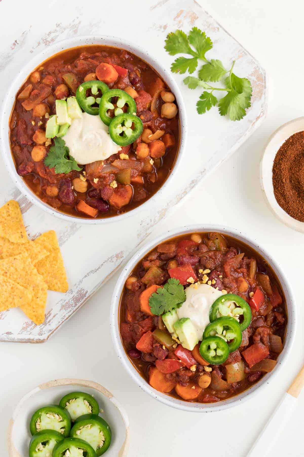 two bowls of chili on a white surface alongside corn tortilla chips, chili powder, cilantro, and sliced jalapeño peppers