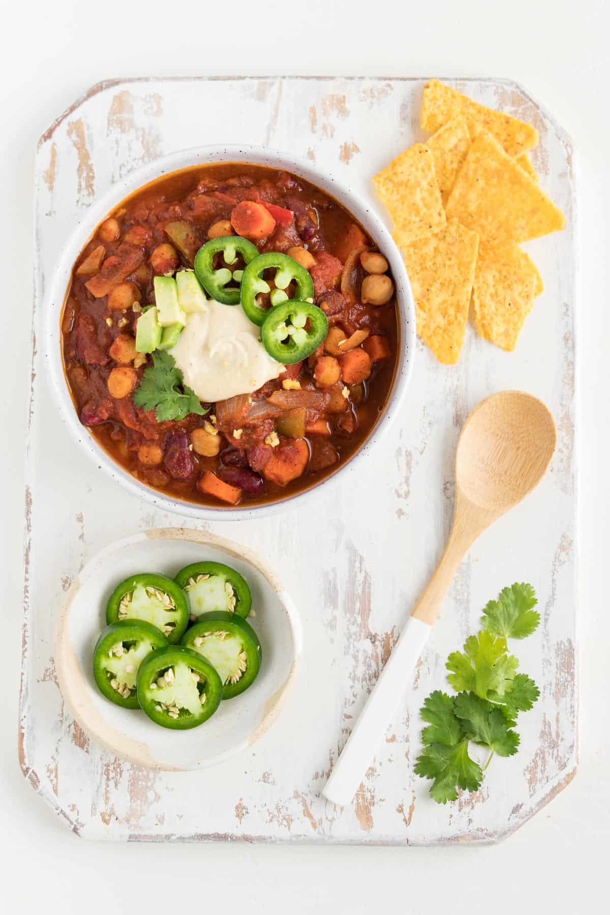 a distressed white wooden board topped with a bowl of three bean chili, corn tortilla chips, a wooden spoon, and sliced jalapeño peppers