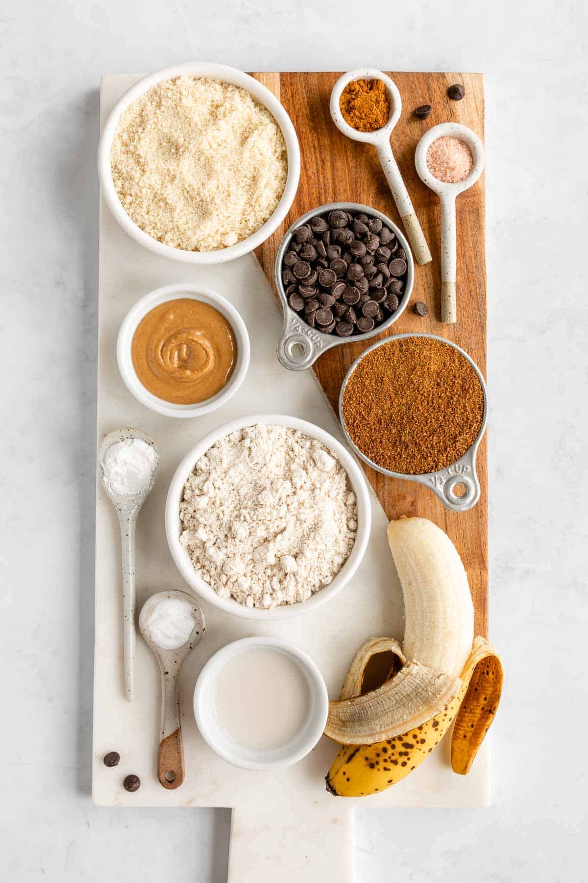 a marble and wood serving board topped with bowls of almond flour, oat flour, coconut sugar, banana, chocolate chips, peanut butter, and almond milk