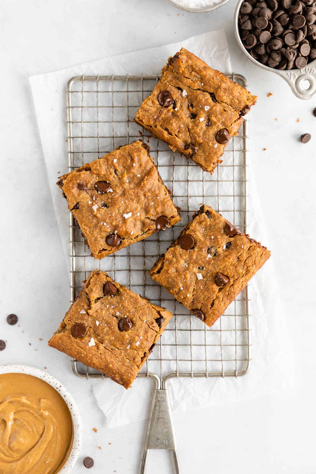 four chocolate chip banana blondies on a wire cooling rack beside a bowl of peanut butter and a cup of chocolate 