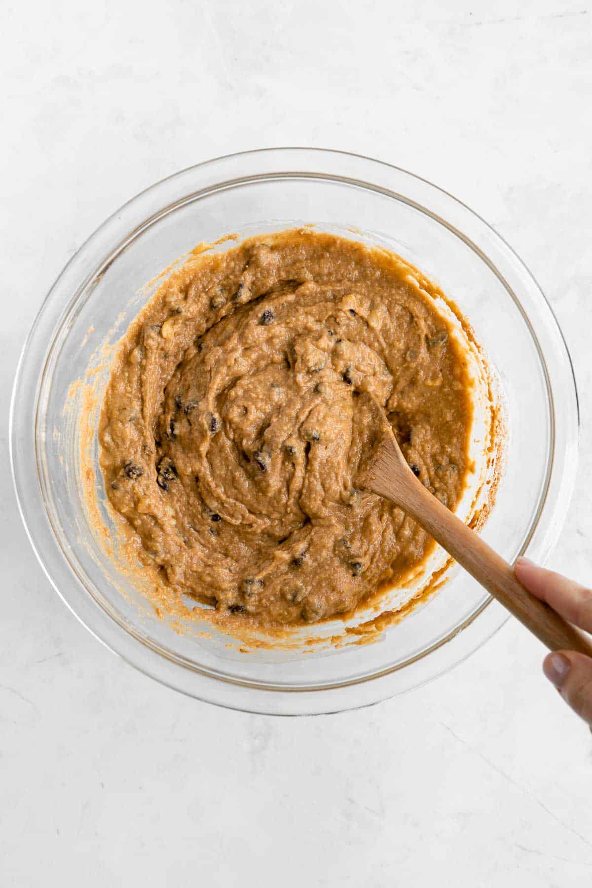 a hand holding a wooden spoon mixing chocolate chip banana bars batter in a glass bowl