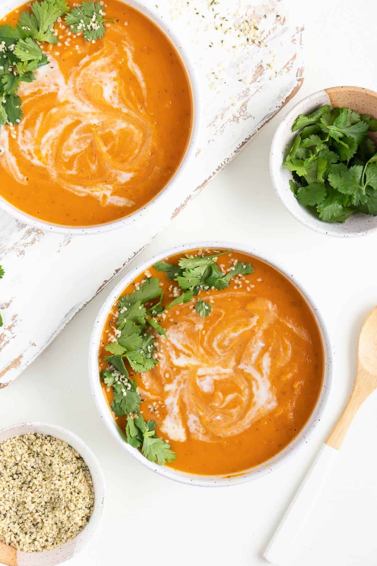 sweet potato soup inside two white bowls beside wooden spoon, a bowl of hemp seeds, and a bowl of cilantro