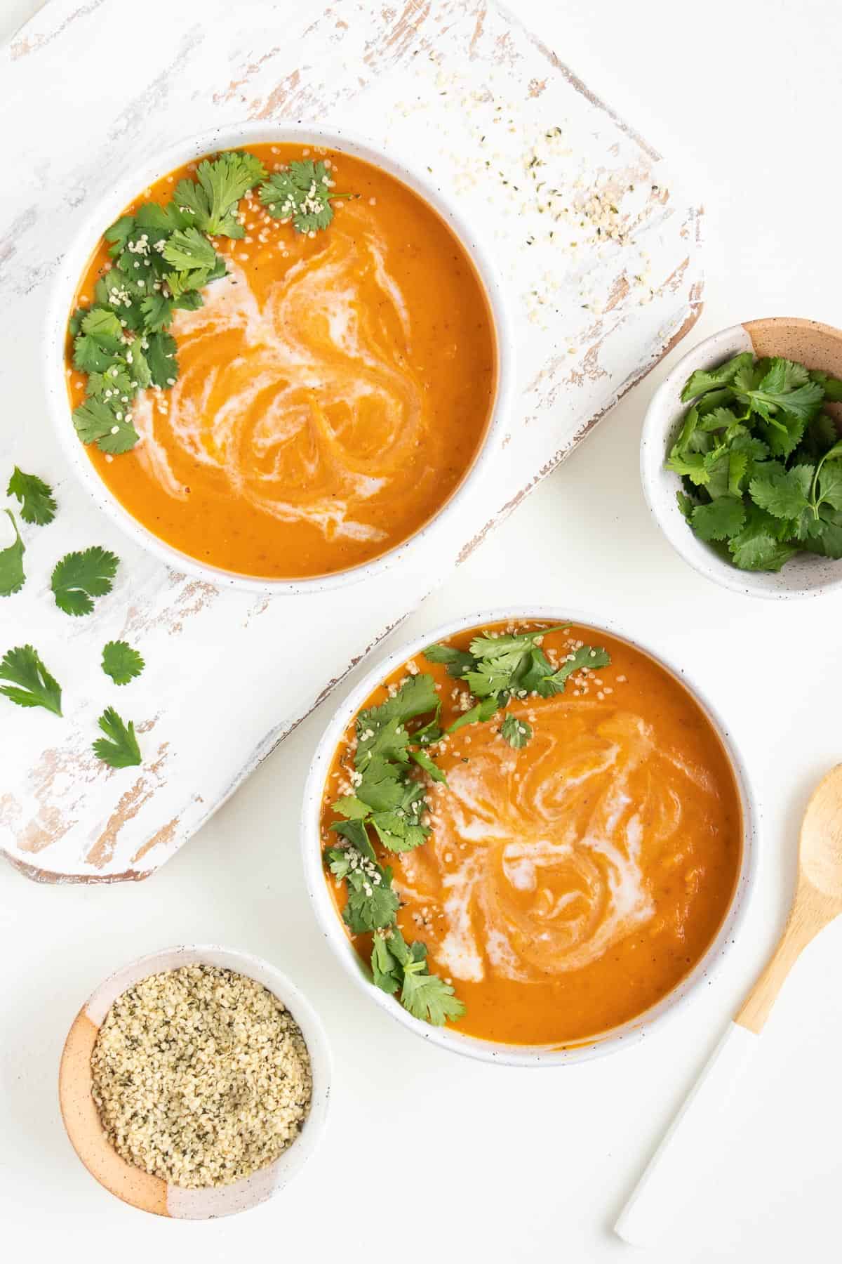 orange soup in two white bowls next to a wooden spoon, a bowl of hemp seeds, and a bowl of cilantro