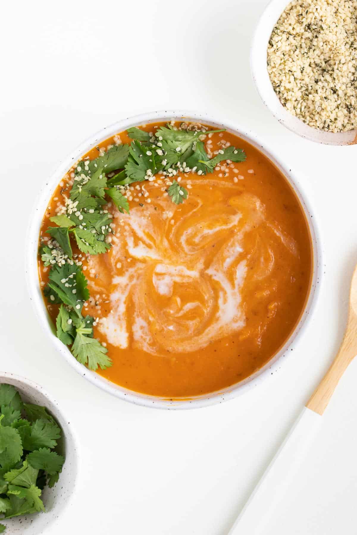 a bowl of creamy vegan sweet potato soup beside a wooden spoon, hemp seeds, and cilantro