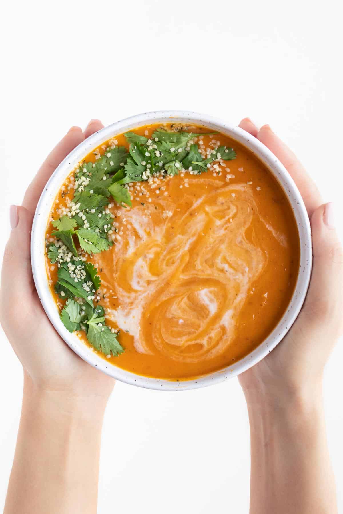 two hands holding a bowl of orange soup with cilantro and hemp seeds on top
