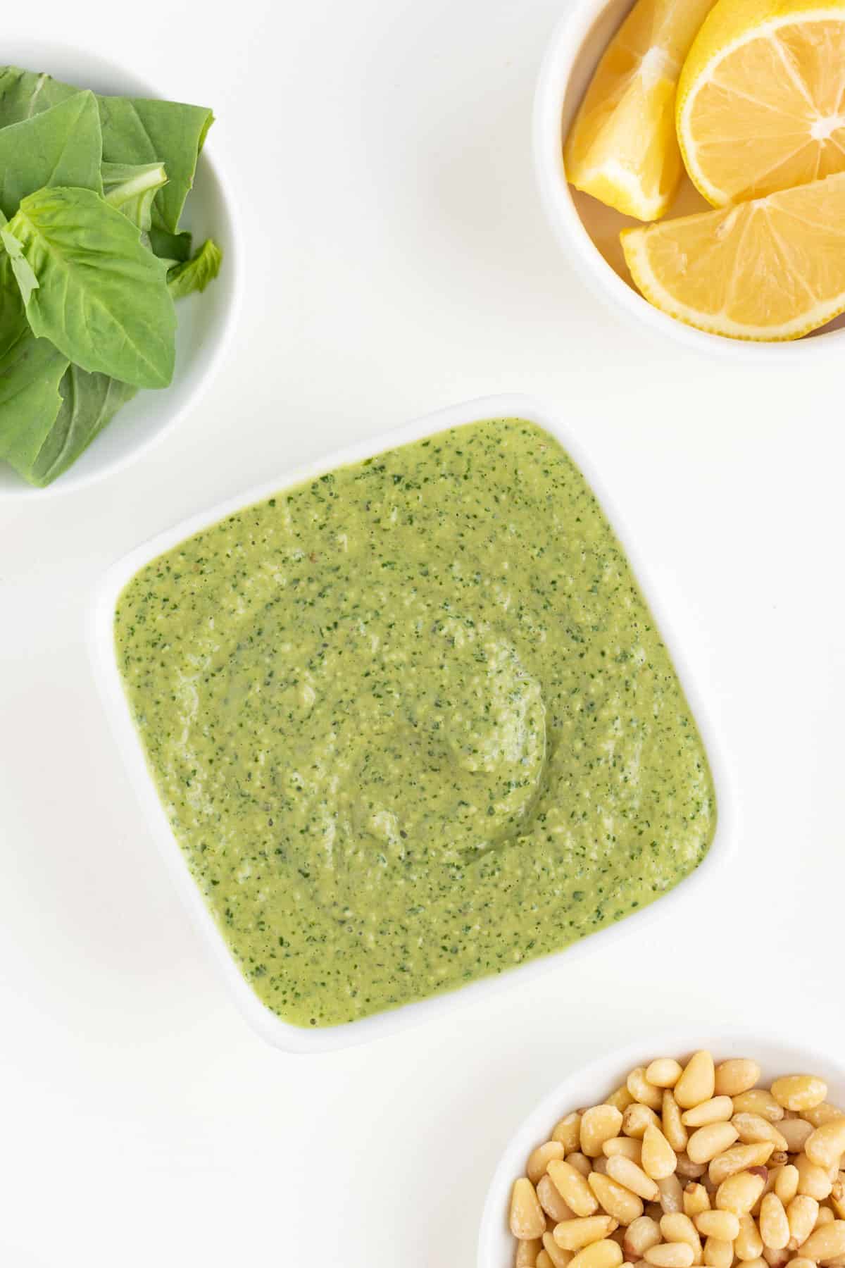 a square white bowl filled with creamy vegan pesto alongside a white bowl filled with pine nuts and a white bowl containing basil leaves