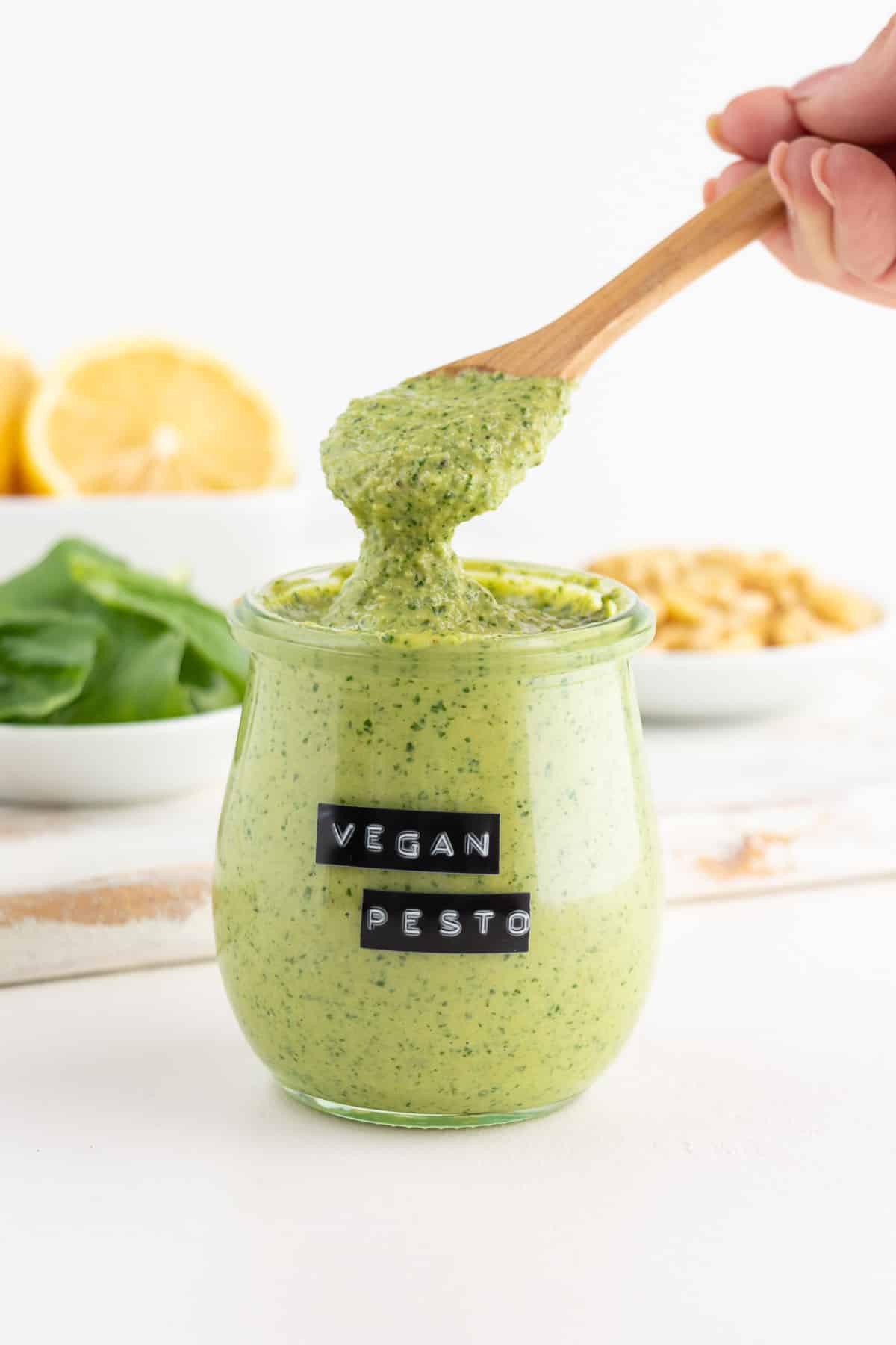 wooden spoon scooping vegan pesto out of a glass jar surrounded by lemons, basil, and pine nuts
