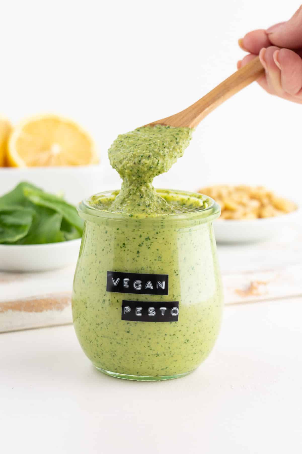wooden spoon scooping vegan pesto out of a glass jar surrounded by lemons, basil, and pine nuts