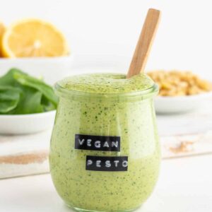 vegan pesto sauce inside a glass jar surrounded by basil leaves, lemon, and pine nuts