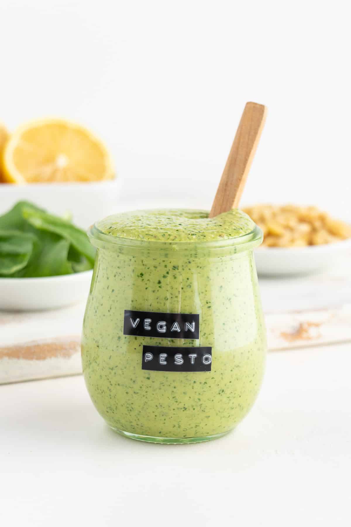 vegan pesto sauce inside a glass jar next to small white bowls filled with basil, pine nuts, and lemons