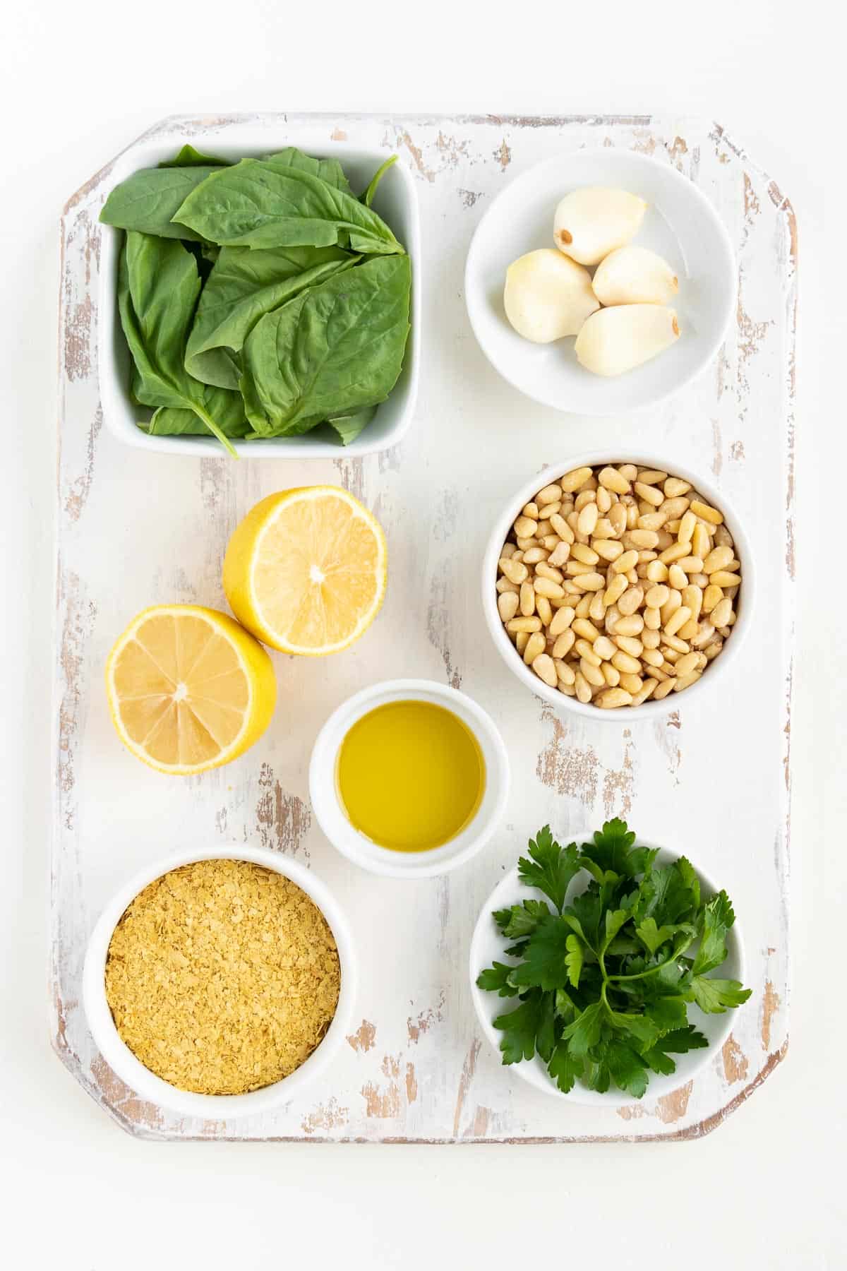 a distressed white wooden cutting board topped with small bowls of basil leaves, garlic cloves, pine nuts, lemon juice, olive oil, nutritional yeast, and parsley