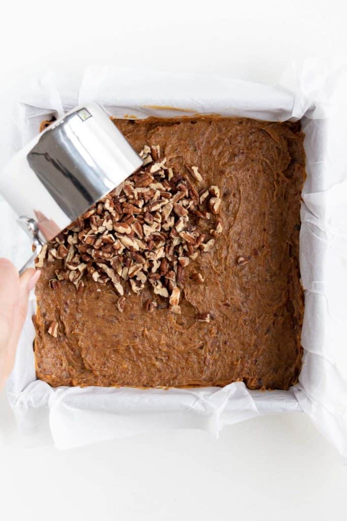 a hand pouring a measuring cup filled with pecans over caramel brownies