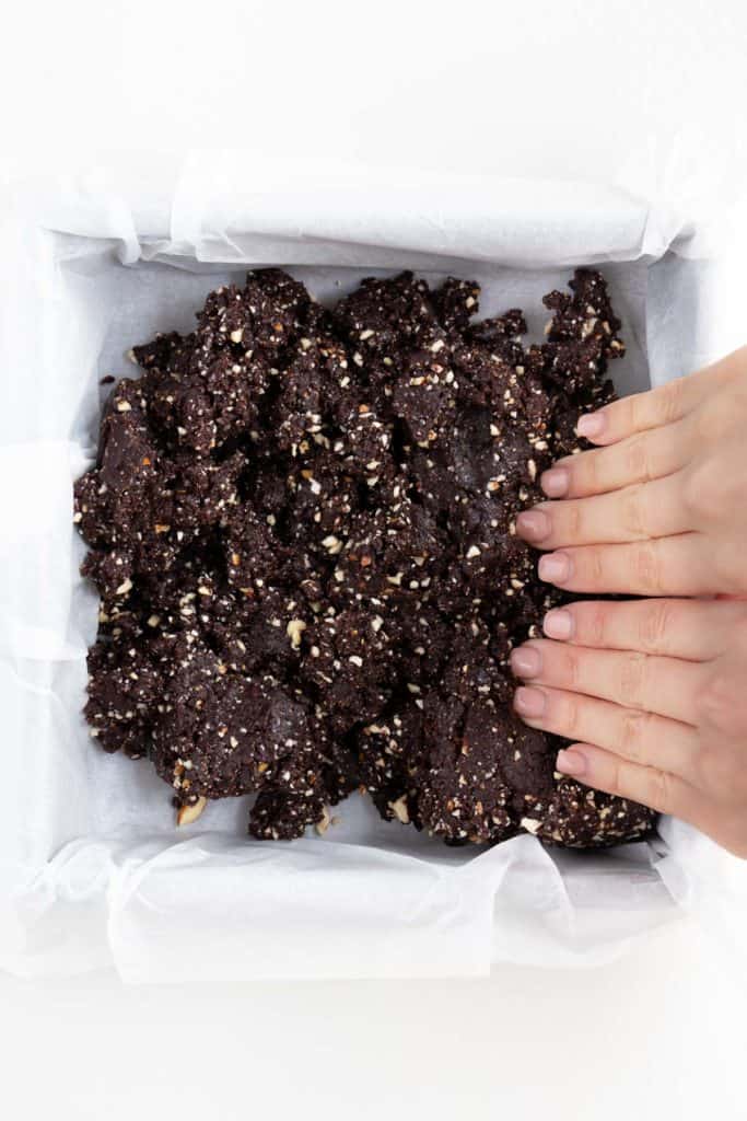 hands pressing raw brownie dough into a square baking pan