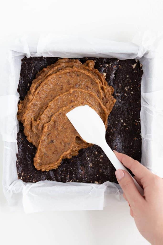 hand holding a white spatula spreading caramel on top of a tray of no-bake brownies