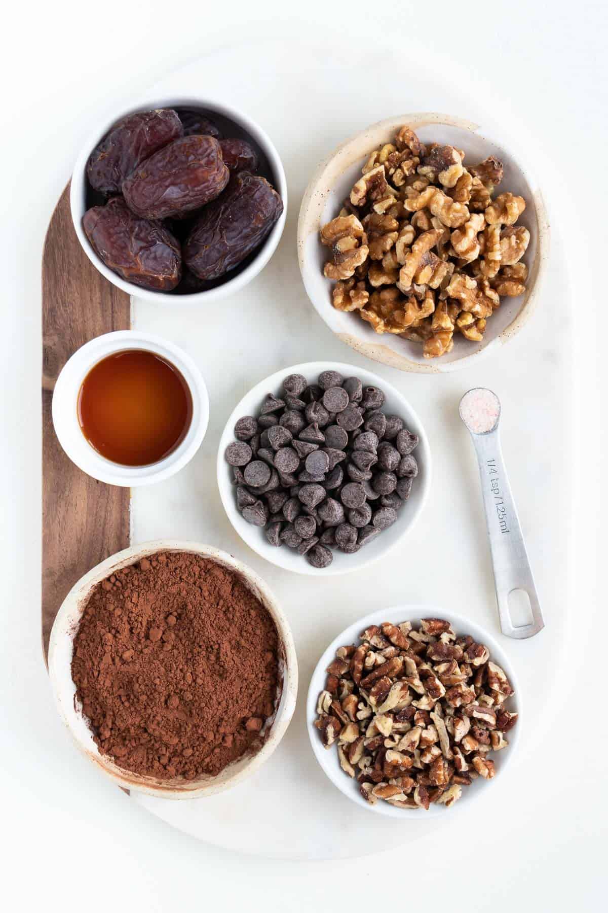 wood and marble cutting board topped with bowls of medjool dates, chocolate chips, walnuts, pecans, maple syrup, and cocoa powder.