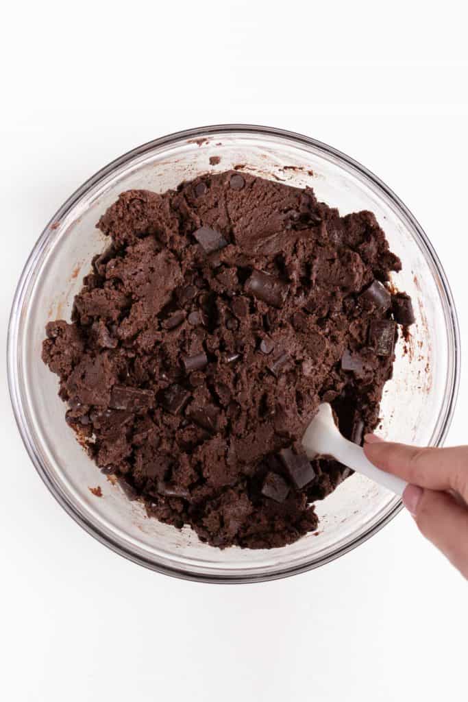 hand holding a white spatula mixing double chocolate chip cookie dough inside a glass bowl
