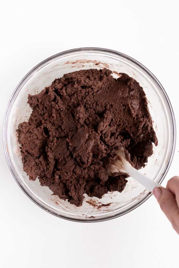 hand holding a white spatula mixing double chocolate cookie dough in a glass bowl