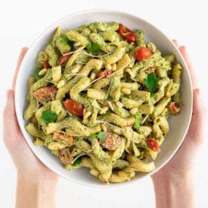 two hands holding a large white bowl of vegan pesto cavatappi pasta