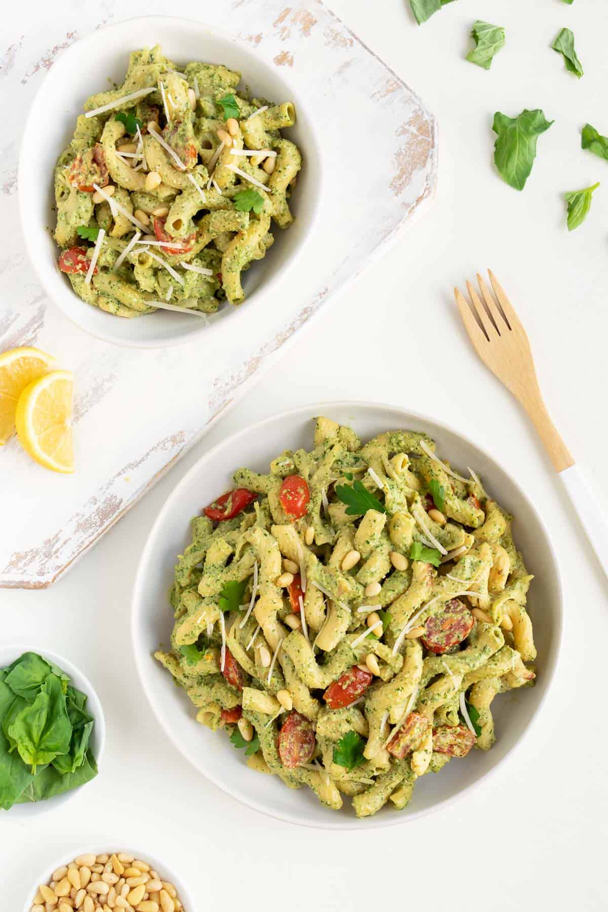 two white bowls filled with vegan pesto cavatappi pasta alongside a small bowl of basil leaves, lemons, a wooden fork, and pine nuts