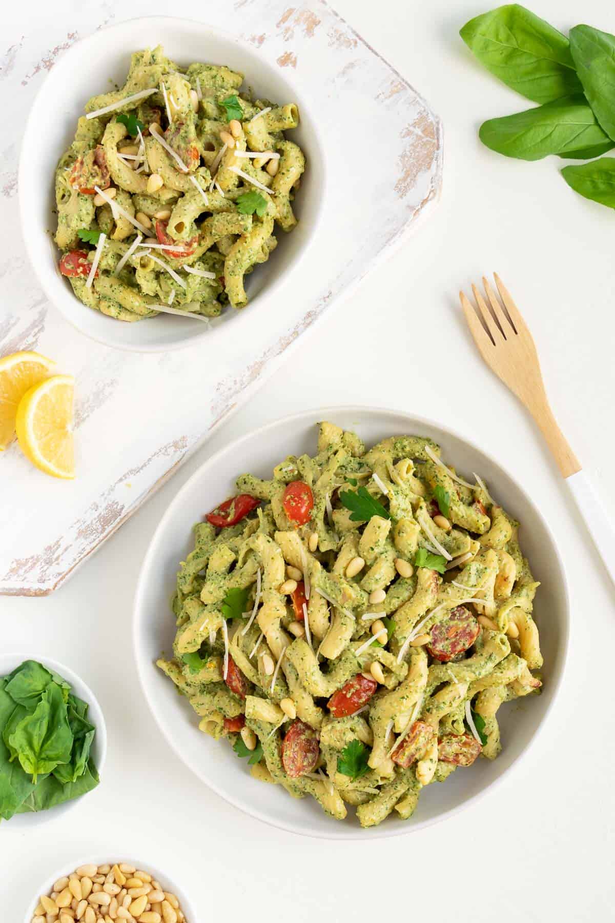 two white bowls filled with vegan pesto cavatappi pasta alongside a small bowl of basil leaves, lemons, a wooden fork, and pine nuts