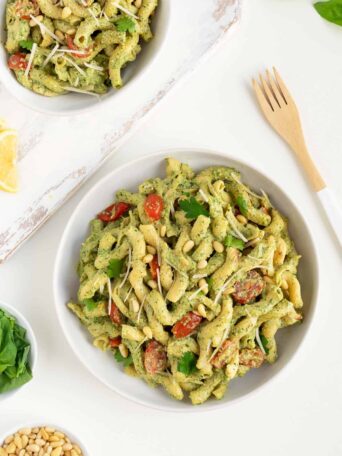 two bowls of vegan pesto cavatappi pasta surrounded by fresh basil, pine nuts, and a wooden fork