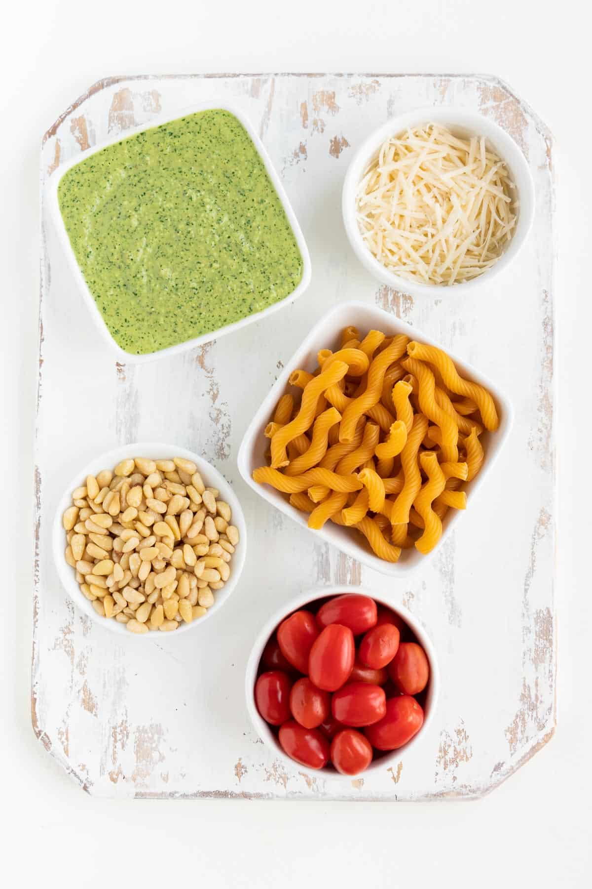 distressed white wood cutting board topped with small bowls of pesto sauce, vegan parmesan cheese, pine nuts, cherry tomatoes, and cavatappi pasta shells