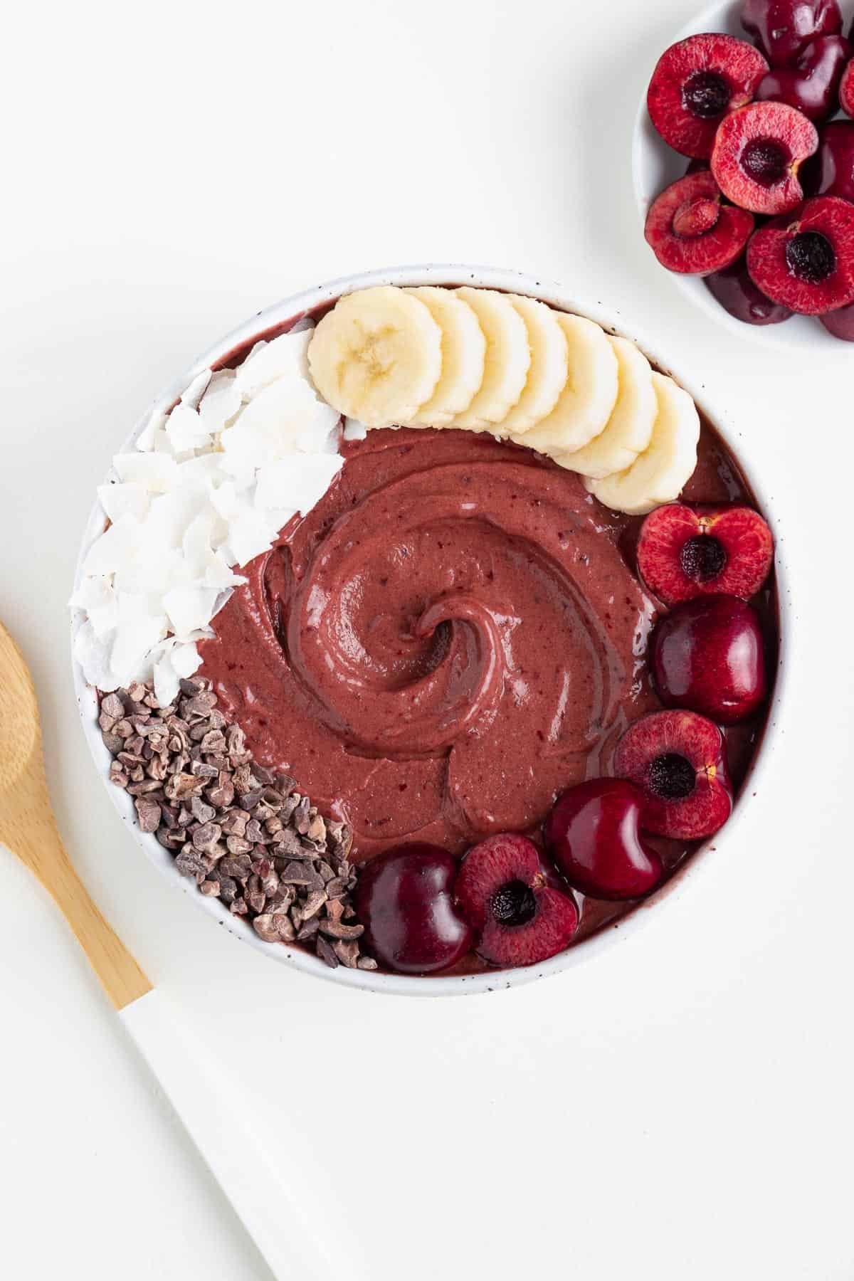 a cherry chocolate smoothie bowl beside a wooden spoon and bowl of black cherries