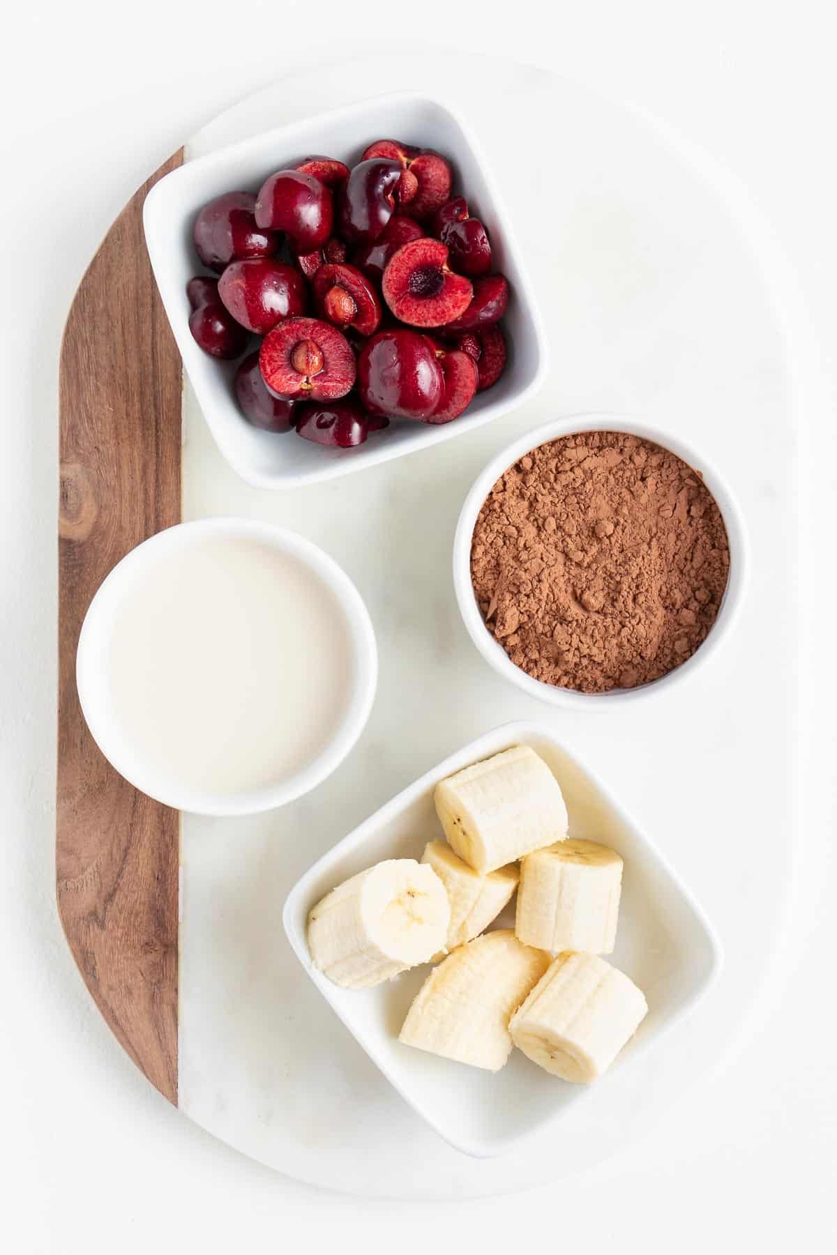 sliced bananas, cocoa powder, almond milk, and berries inside bowls on a white marble board