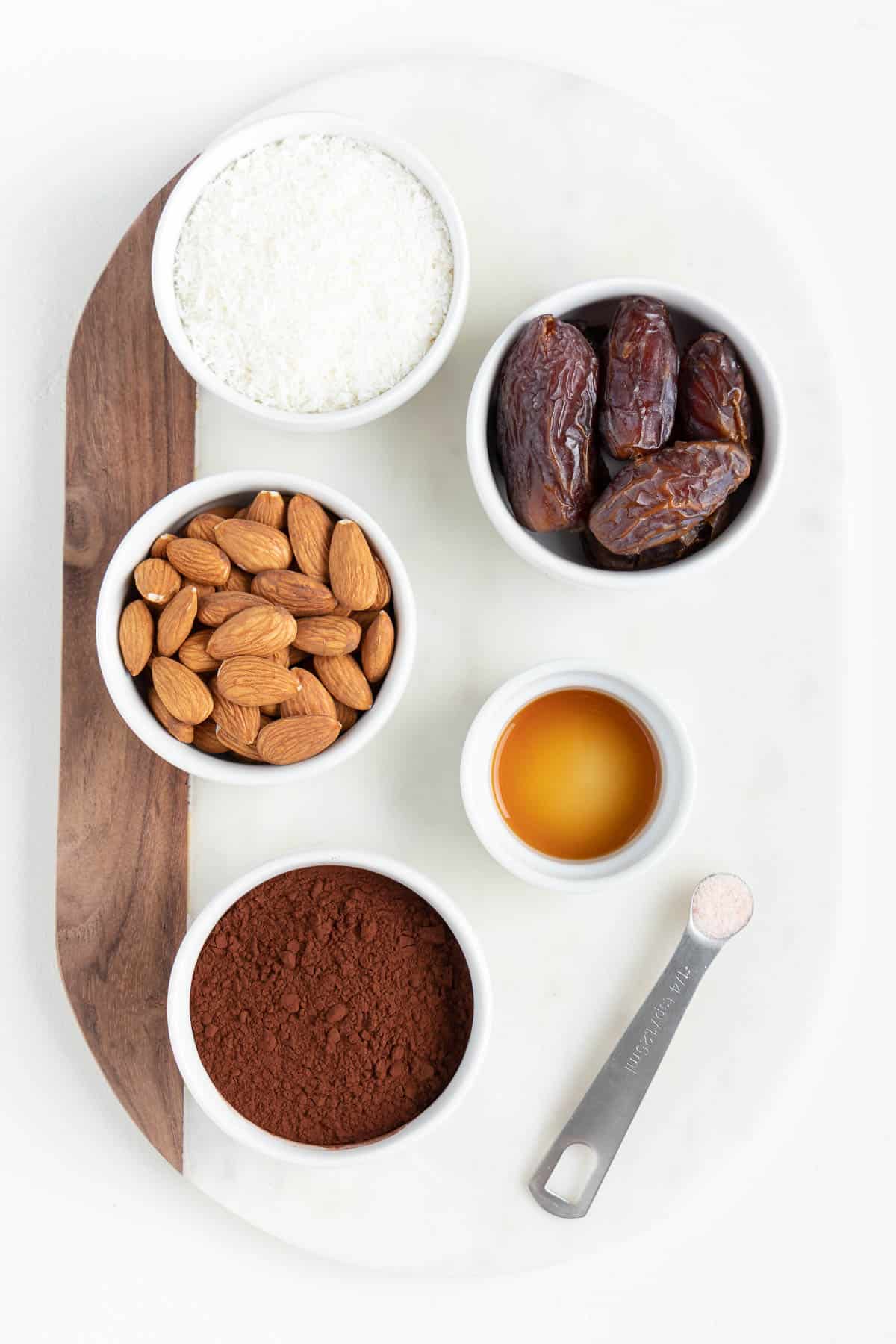 small white bowls filled with raw almonds, medjool dates, cacao powder, vanilla extract, and a teaspoon of salt on a marble board