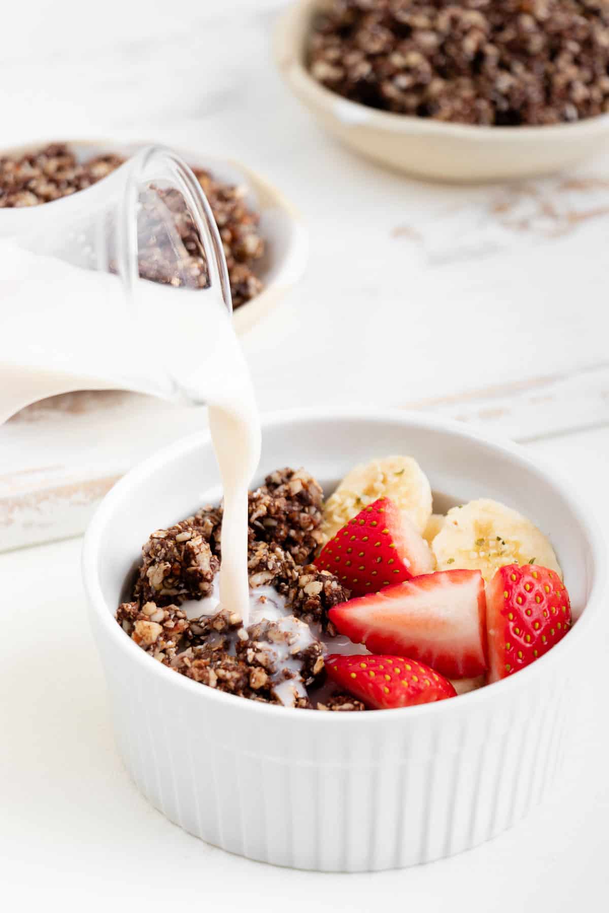 almond milk being poured over a bowl of chocolate rawnola with strawberries and banana