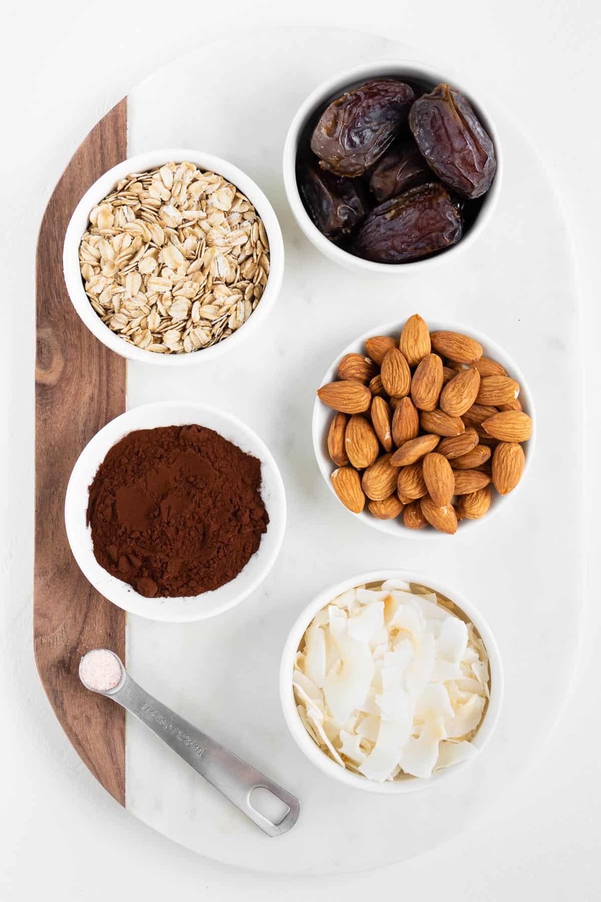 white bowls filled with rolled oats, almonds, medjool dates, cocoa powder, and coconut flakes