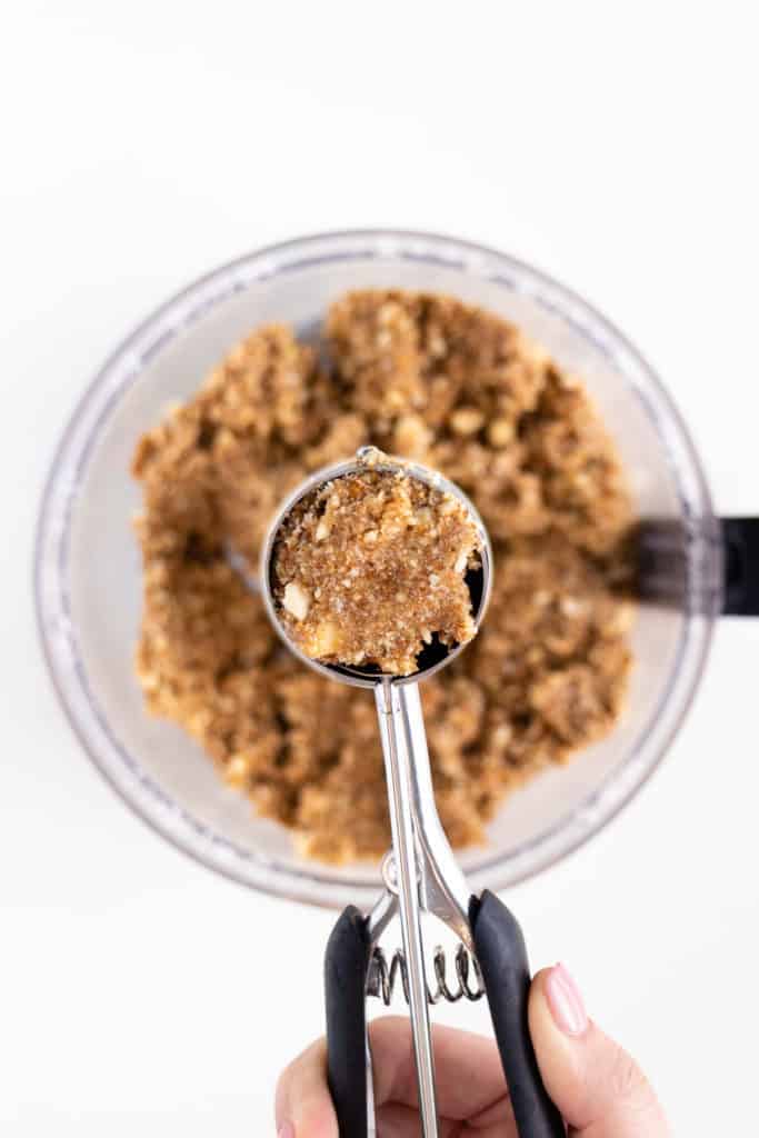 hand holding a cookie scoop filled with dough in front of a food processor