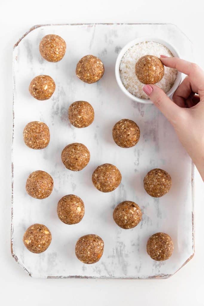 a hand rolling a ball of no-bake dough into a bowl of shredded coconut surrounded by several energy balls