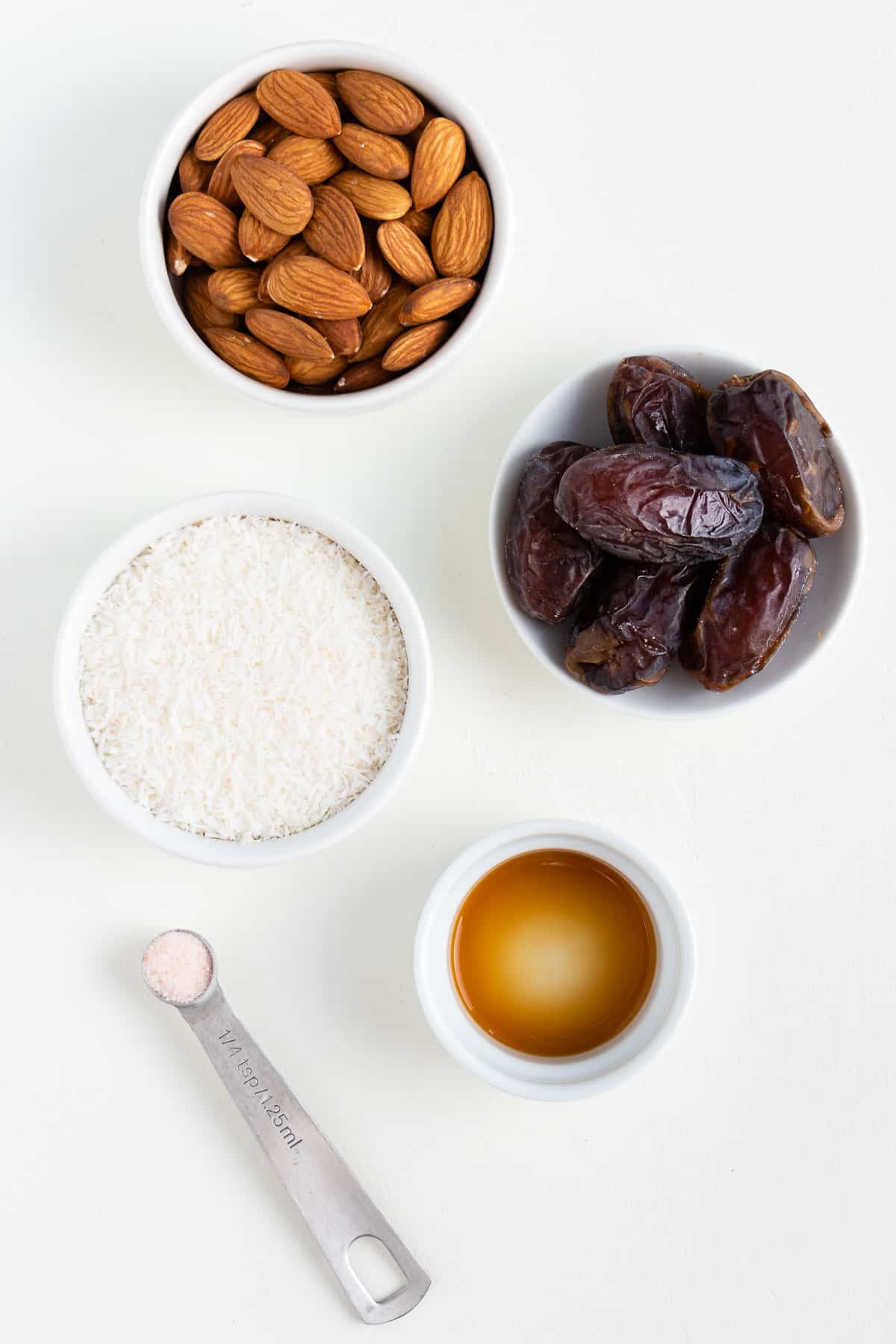 small white bowls of nuts, coconut, dates, and vanilla extract on a white surface