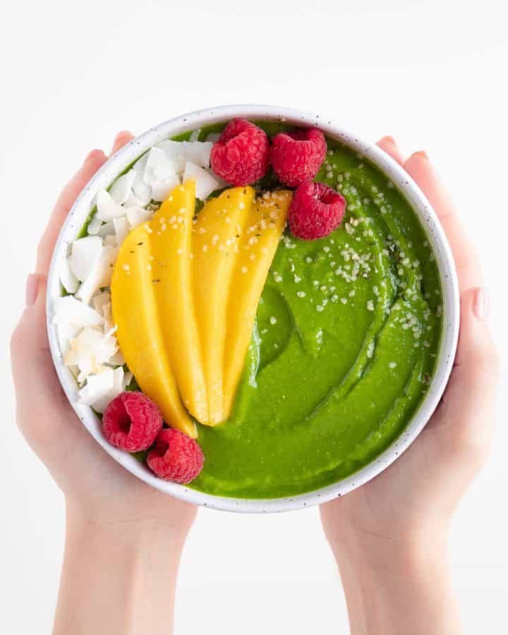 two hands holding a green smoothie bowl topped with sliced mango, coconut flakes, raspberries, and hemp seeds