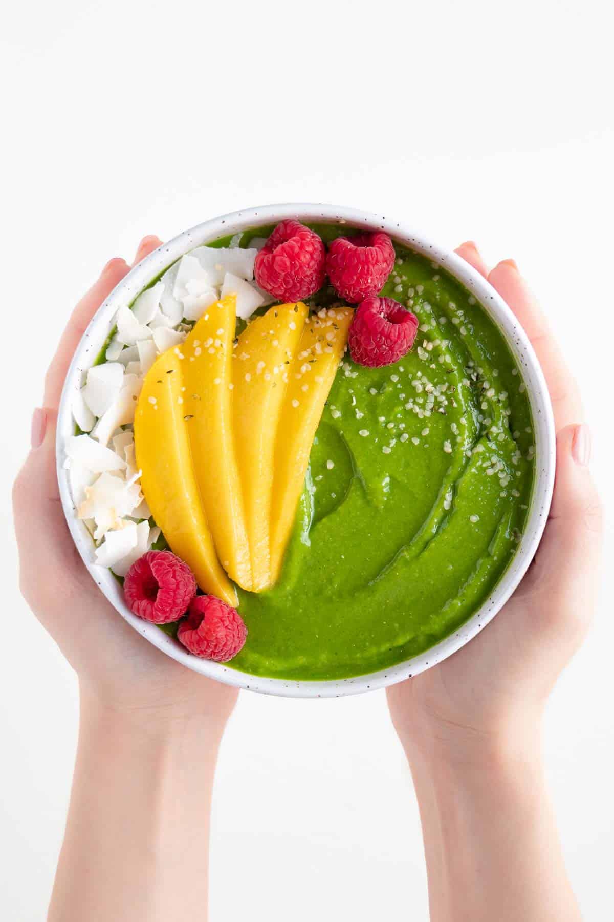 two hands holding a green smoothie bowl topped with sliced mango, coconut flakes, raspberries, and hemp seeds