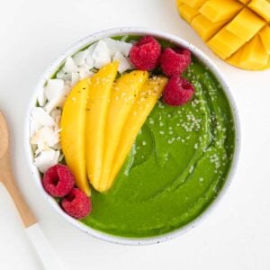 a bowl filled with a green smoothie and fresh fruit beside a wooden spoon