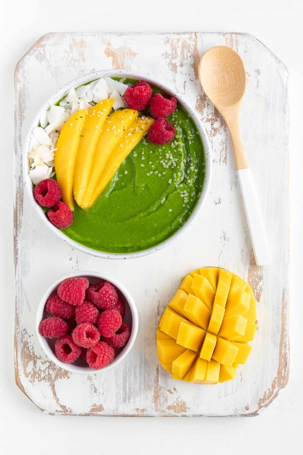 green mango smoothie bowl, sliced mango, and a bowl of raspberries on top of a distressed cutting board