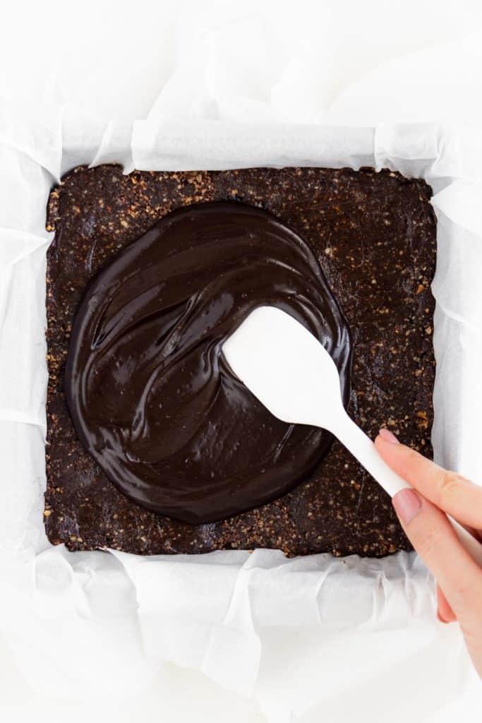 a hand holding a white spatula spreading chocolate ganache over no bake vegan brownies