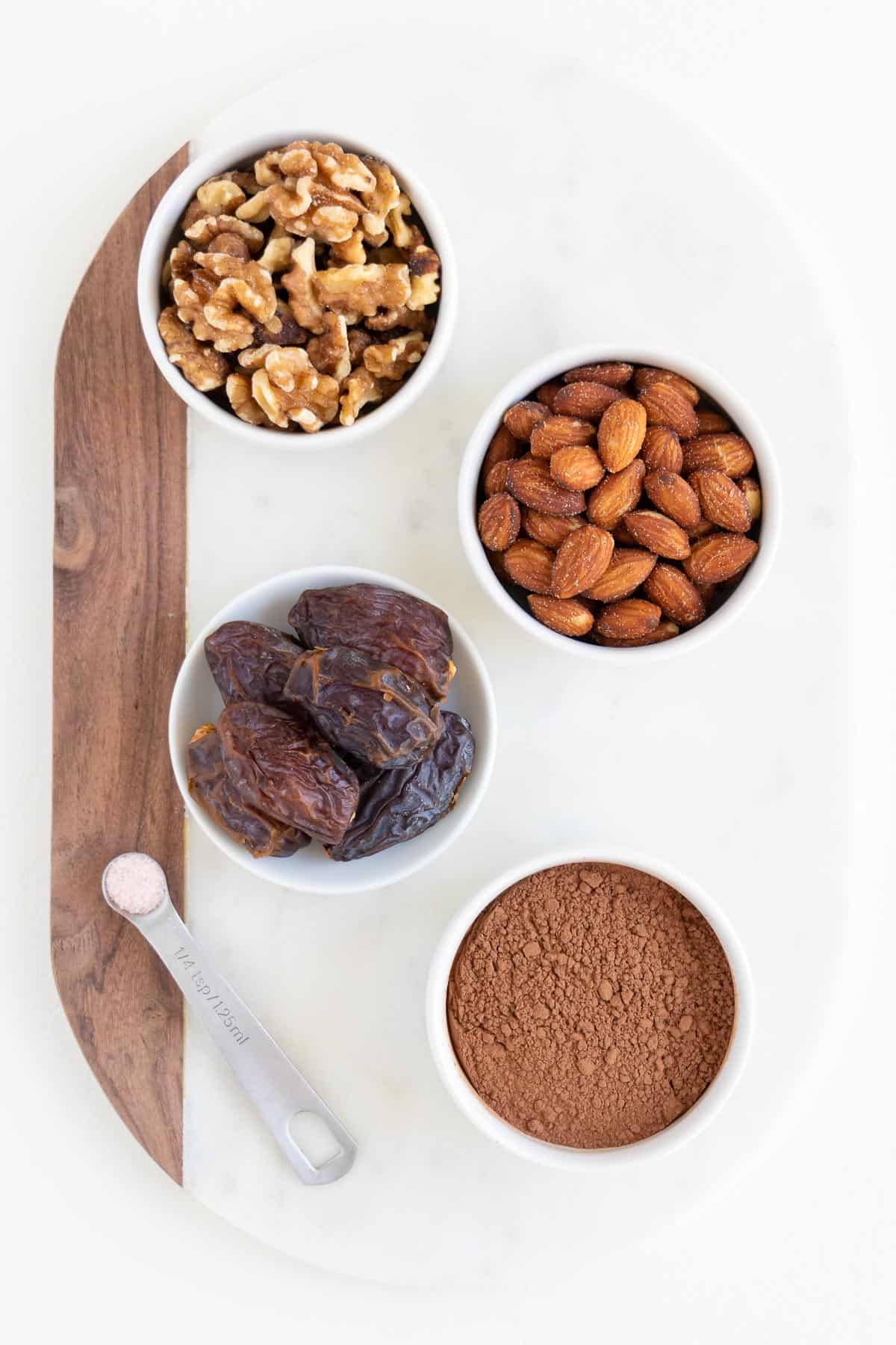 white bowls filled with walnuts, almonds, dates, and cacao powder on a white marble cutting board