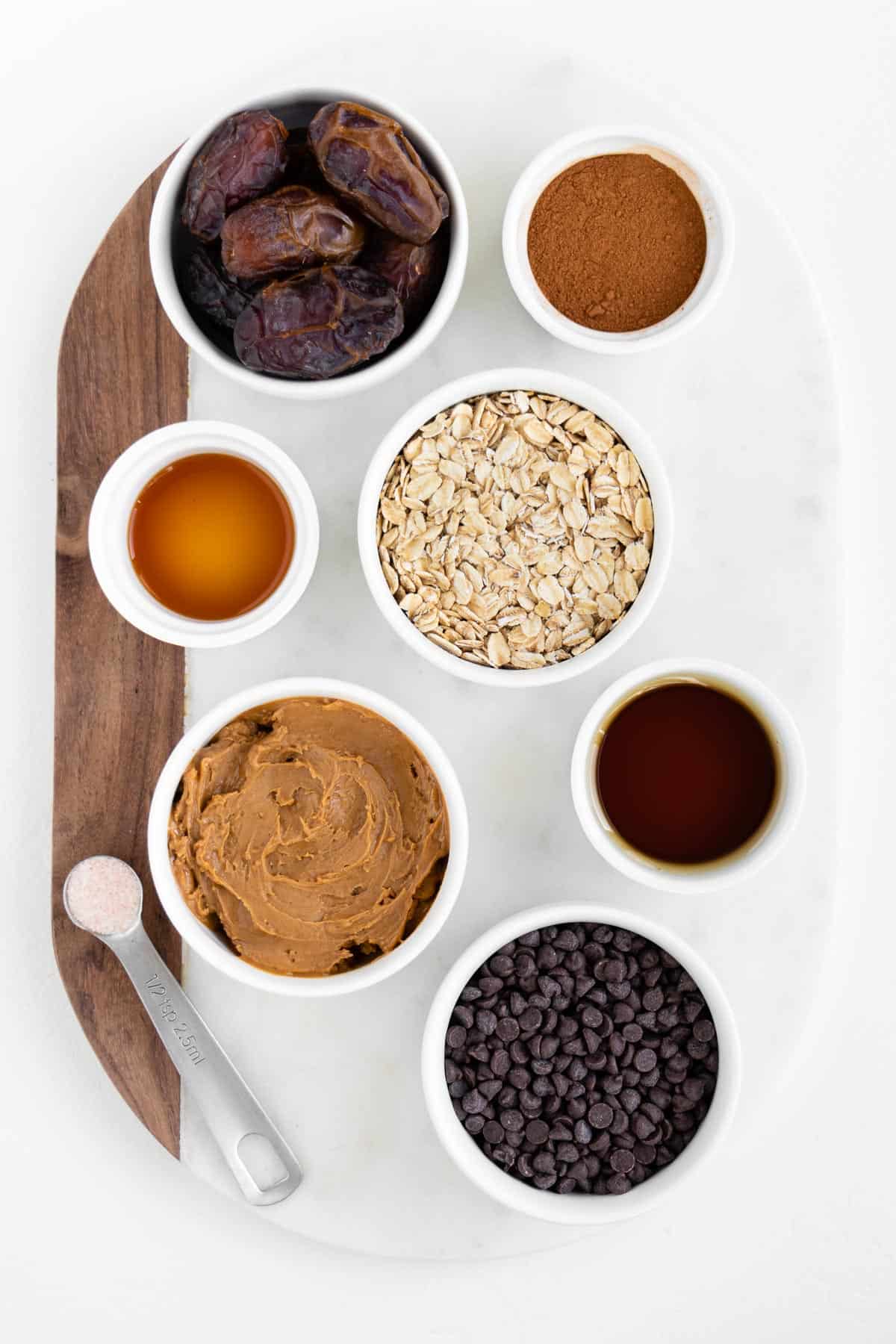 small white bowls of medjool dates, peanut butter, cinnamon, vanilla, chocolate chips, maple syrup, and salt on a white marble board